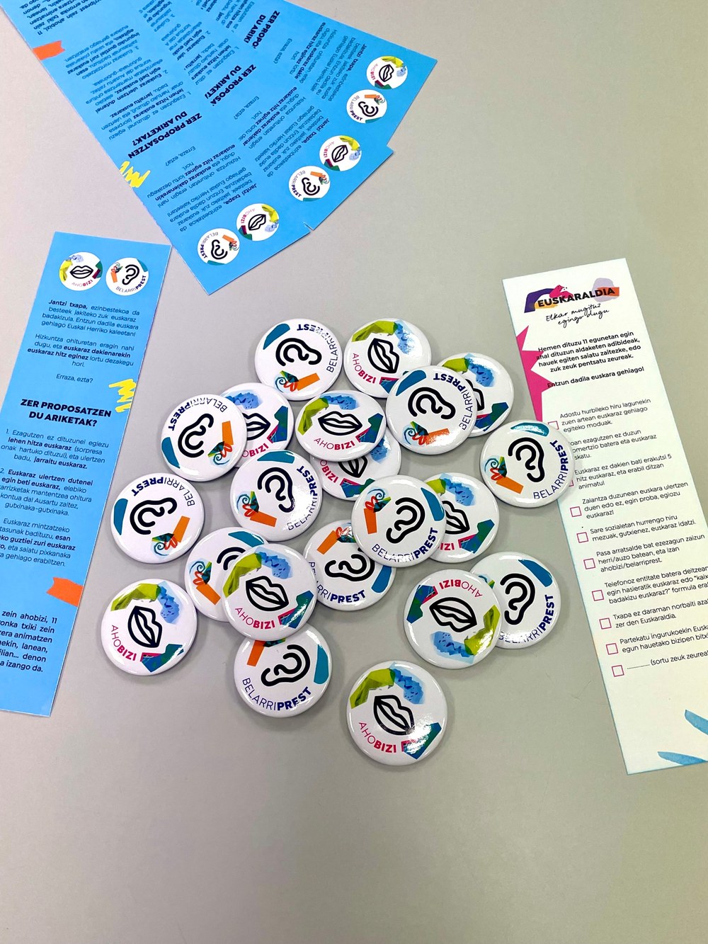 Chapas Ahobizi y Belarriprest del Euskaraldia de 2025, junto a los marcapáginas que se repartirán con las chapas.