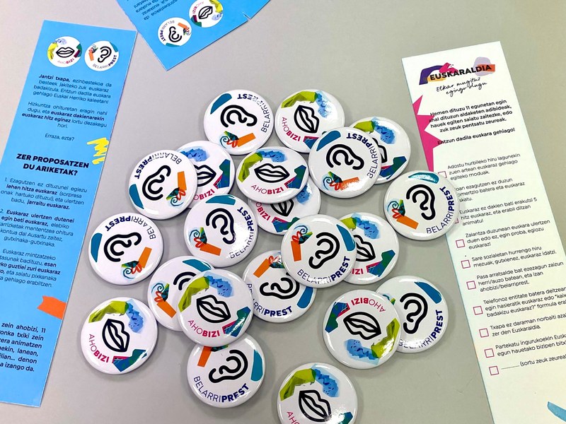 Chapas Ahobizi y Belarriprest del Euskaraldia de 2025, junto a los marcapáginas que se repartirán con las chapas.